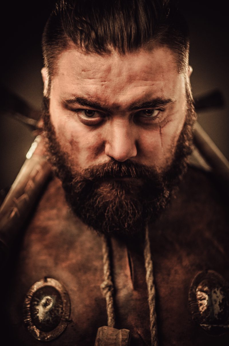 Serious viking with sword in a traditional warrior clothes, posing on a dark background.